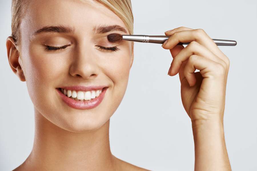 Femme souriante appliquant une astuce beauté de maquillage rapide avec un pinceau sur sa paupière.