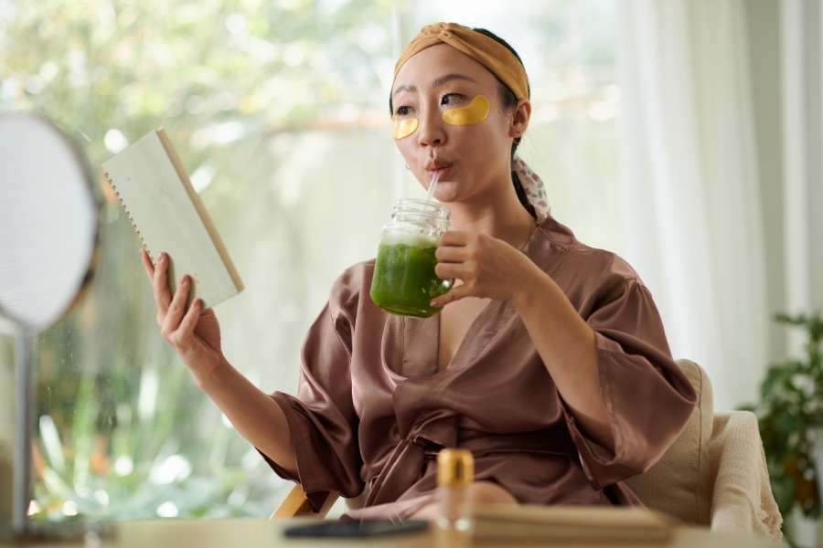 Femme buvant un smoothie vert avec masque facial dans sa routine matinale énergisante