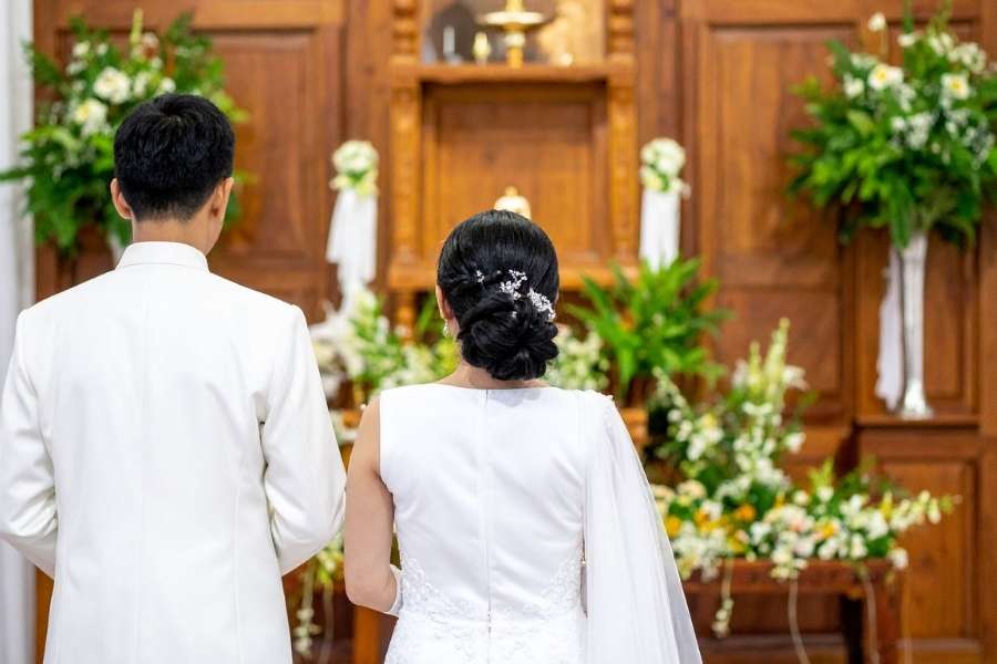 Vue de dos d'un couple en tenue de mariage blanche debout devant un autel en bois traditionnel richement décoré de compositions florales lors d'une cérémonie religieuse.