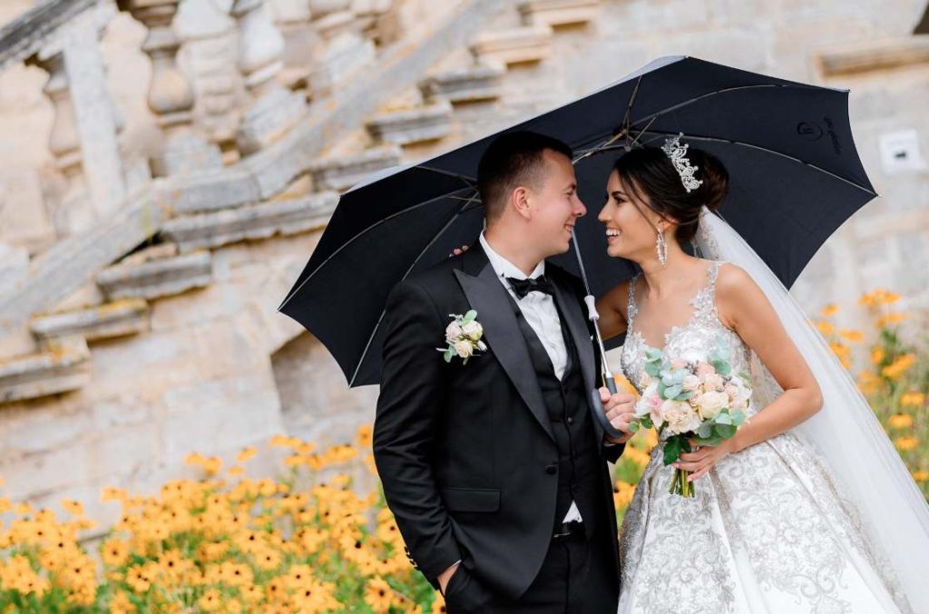 Couple de mariés souriant sous un parapluie noir dans un jardin, illustrant comment surmonter les principales erreurs organisation mariage liées à la météo