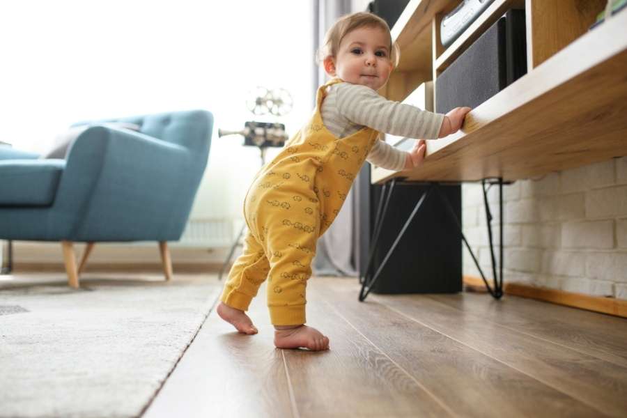 Un bébé en combinaison jaune apprend à se mettre debout en s'appuyant sur un meuble bas dans un salon avant de faire ses premiers pas.